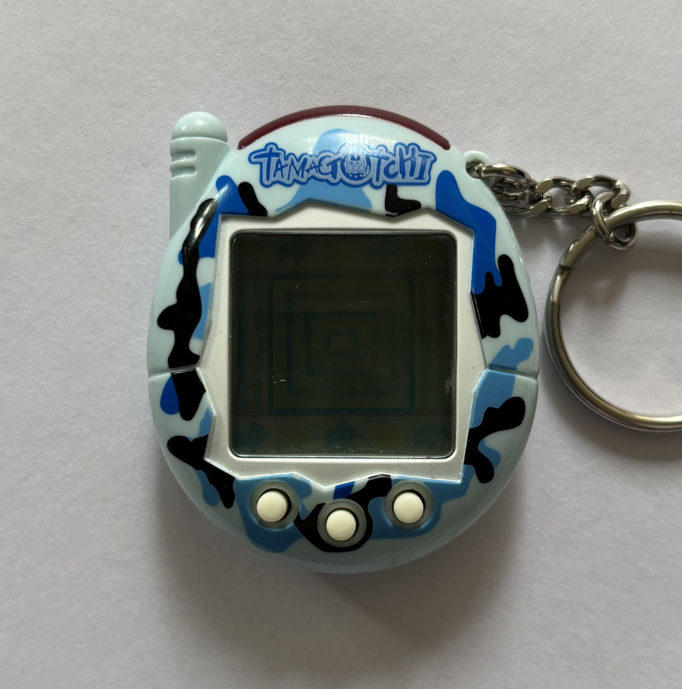 Light blue shell. White screen border and buttons. Blue/black camo print surrounding screen. 'Tamagotchi' label on blue background.