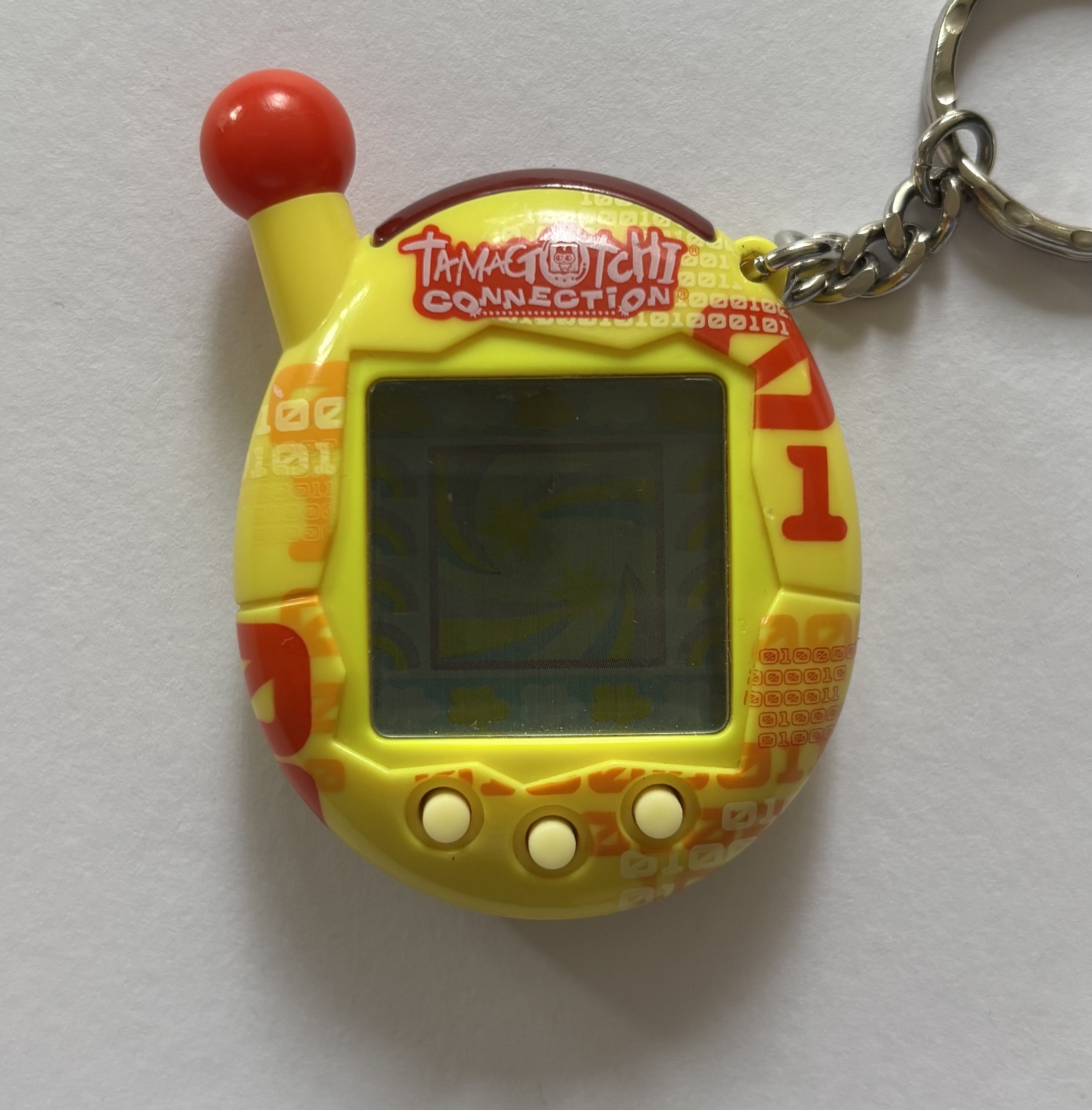 Yellow shell, screen border, buttons. Red antenna ball. Orange/yellow/red binary print across front. 'Tamagotchi Connection' on red background.