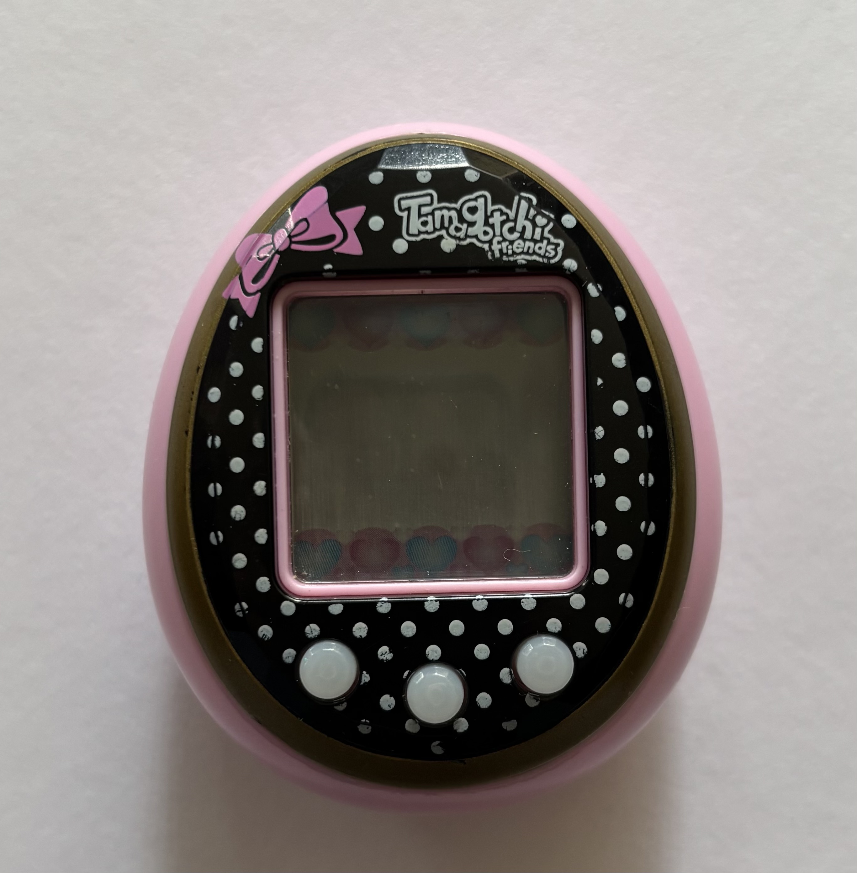 Pink shell. Black faceplate with white polkadots motif. White buttons. Pink bow above screen. 'Tamagotchi Friends' label.