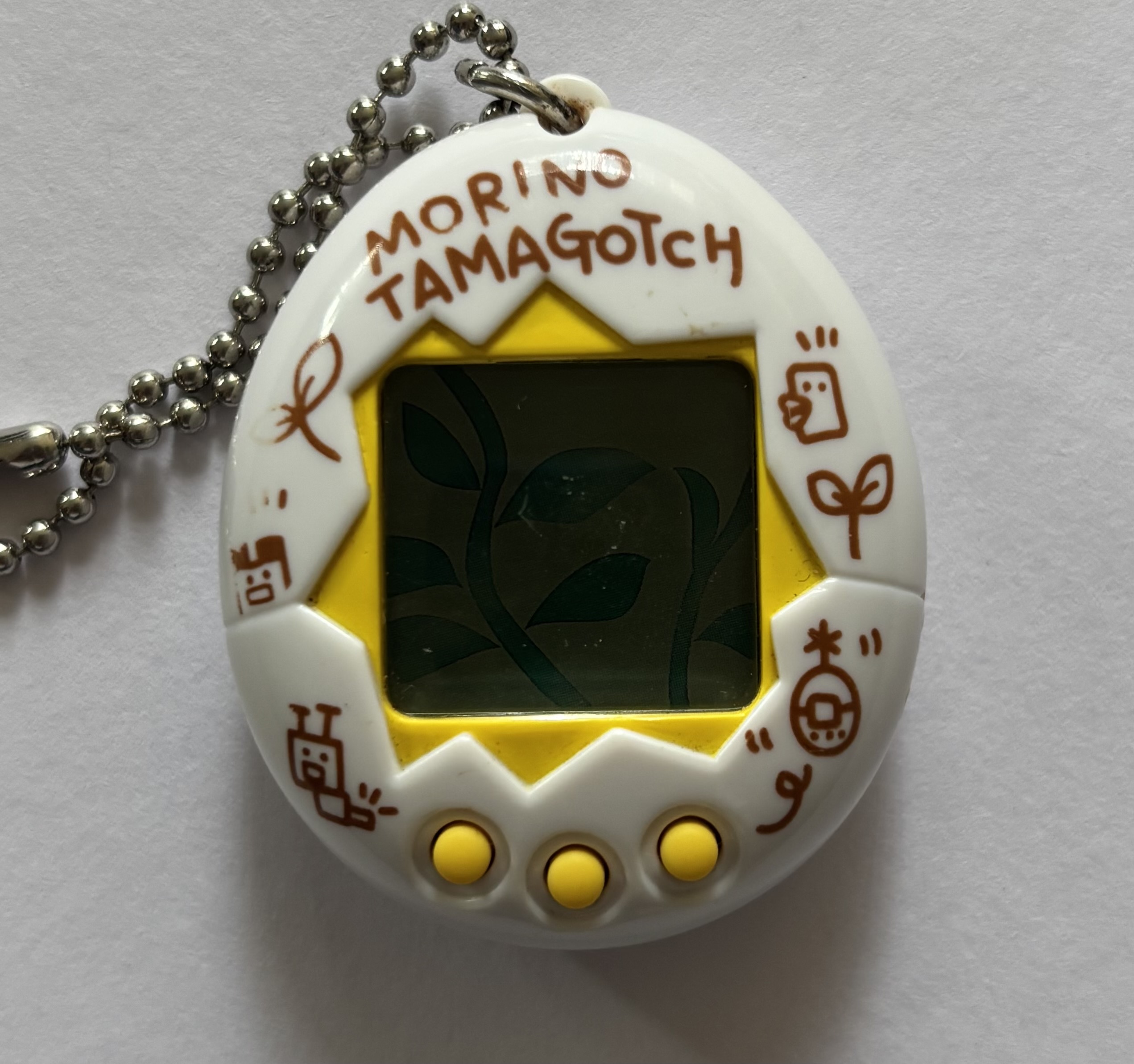 White shell. Yellow screen border and buttons. Brown 'Morino Tamagotch' label and leaf/bug characters surrounding screen.