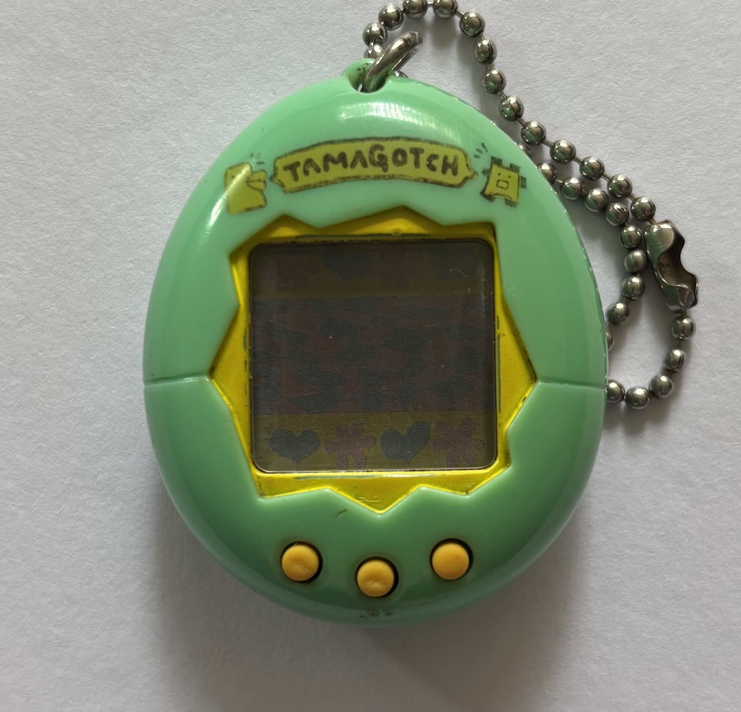 Light blue green shell. Yellow screen border and buttons, 'tamagotch' banner, and characters above screen.