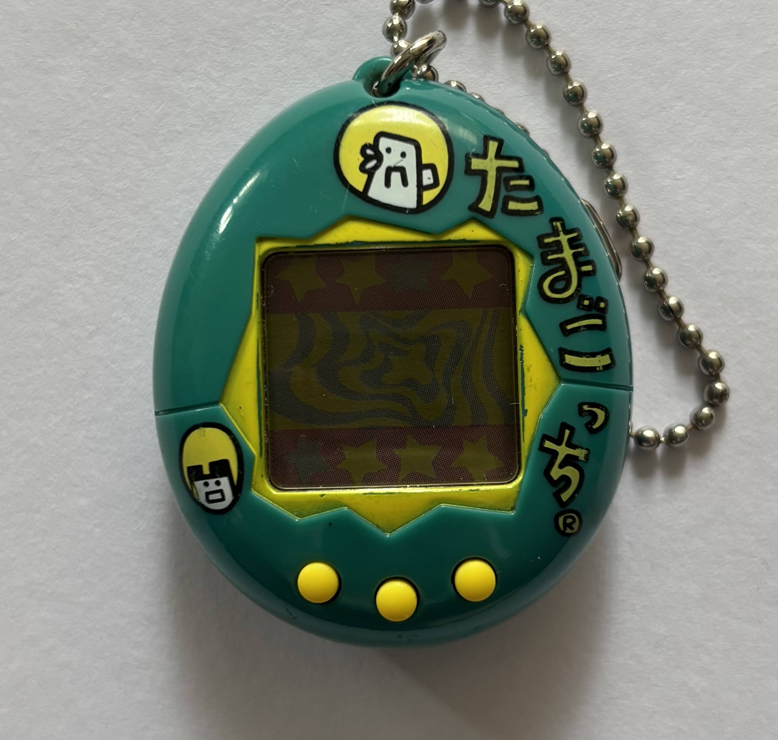 Dark blue green shell. Yellow screen border and buttons. Yellow 'tamagotchi' label in Japanese writing and white characters on yellow backgrounds surrounding screen.