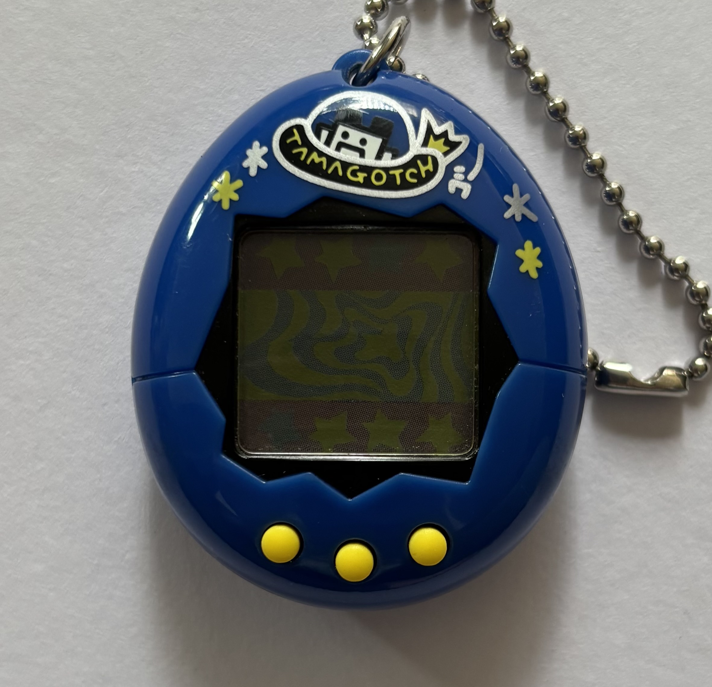 Dark blue shell. Black screen border. Yellow buttons. Character in UFO spaceship labelled 'tamagotch', surrounded by yellow and silver stars.