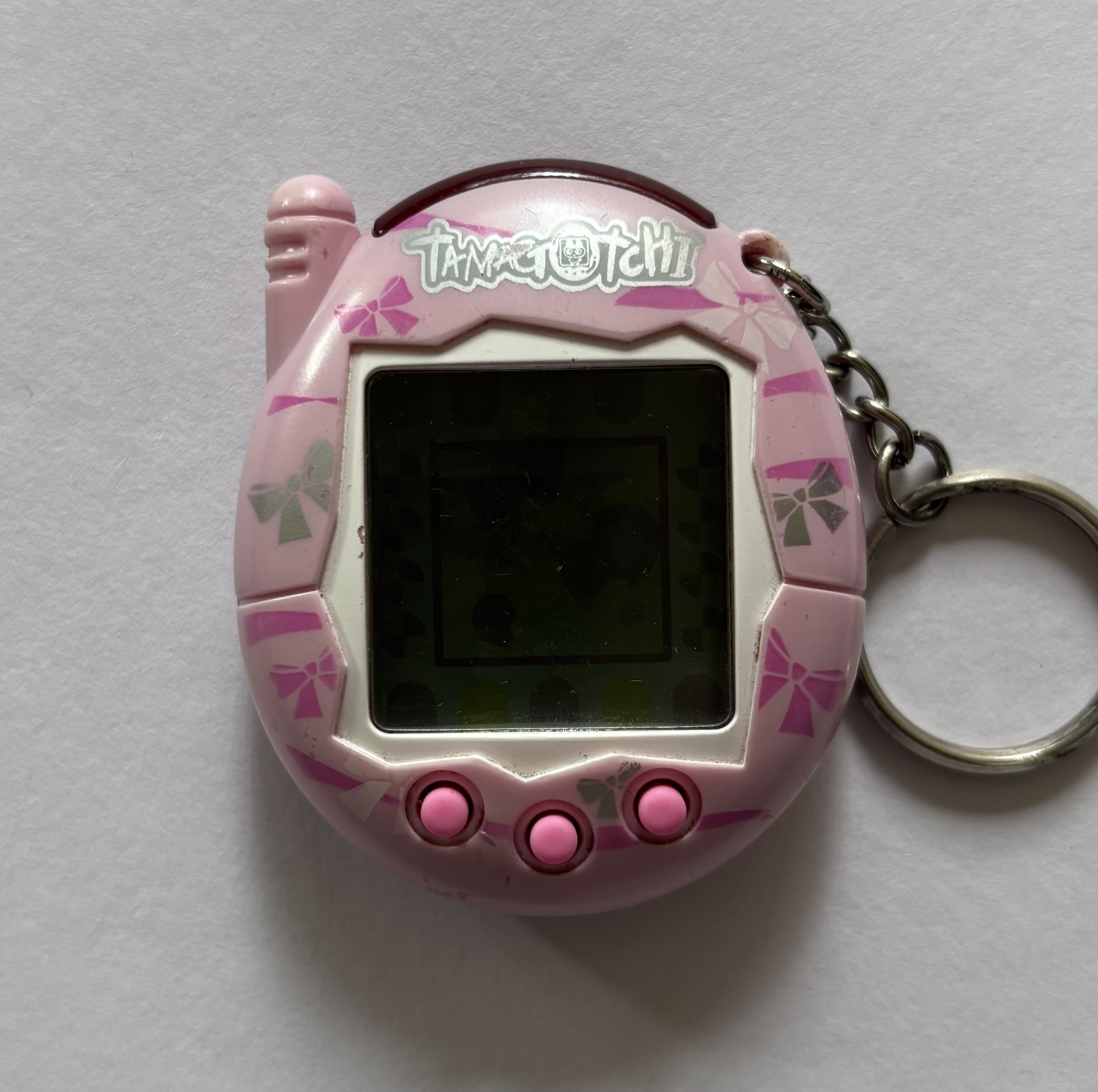Light pink shell. White screen border. Pink buttons. Pink/silver bows surrounding screen. Silver 'Tamagotchi' label.