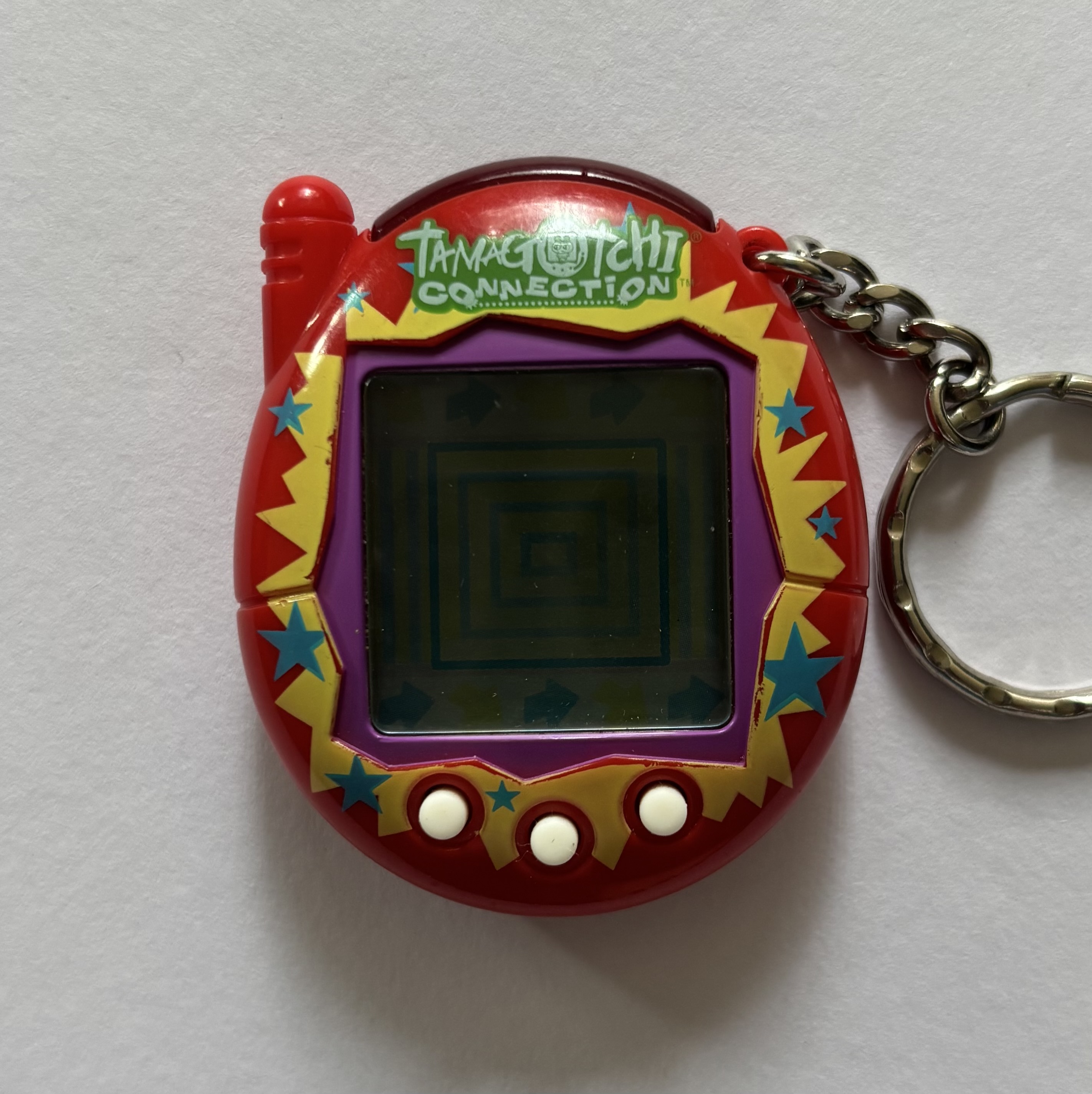Red shell. Purple screen border. White buttons. Yellow sunburst, blue stars surrounding screen. 'Tamagotchi Connection' label on green background.