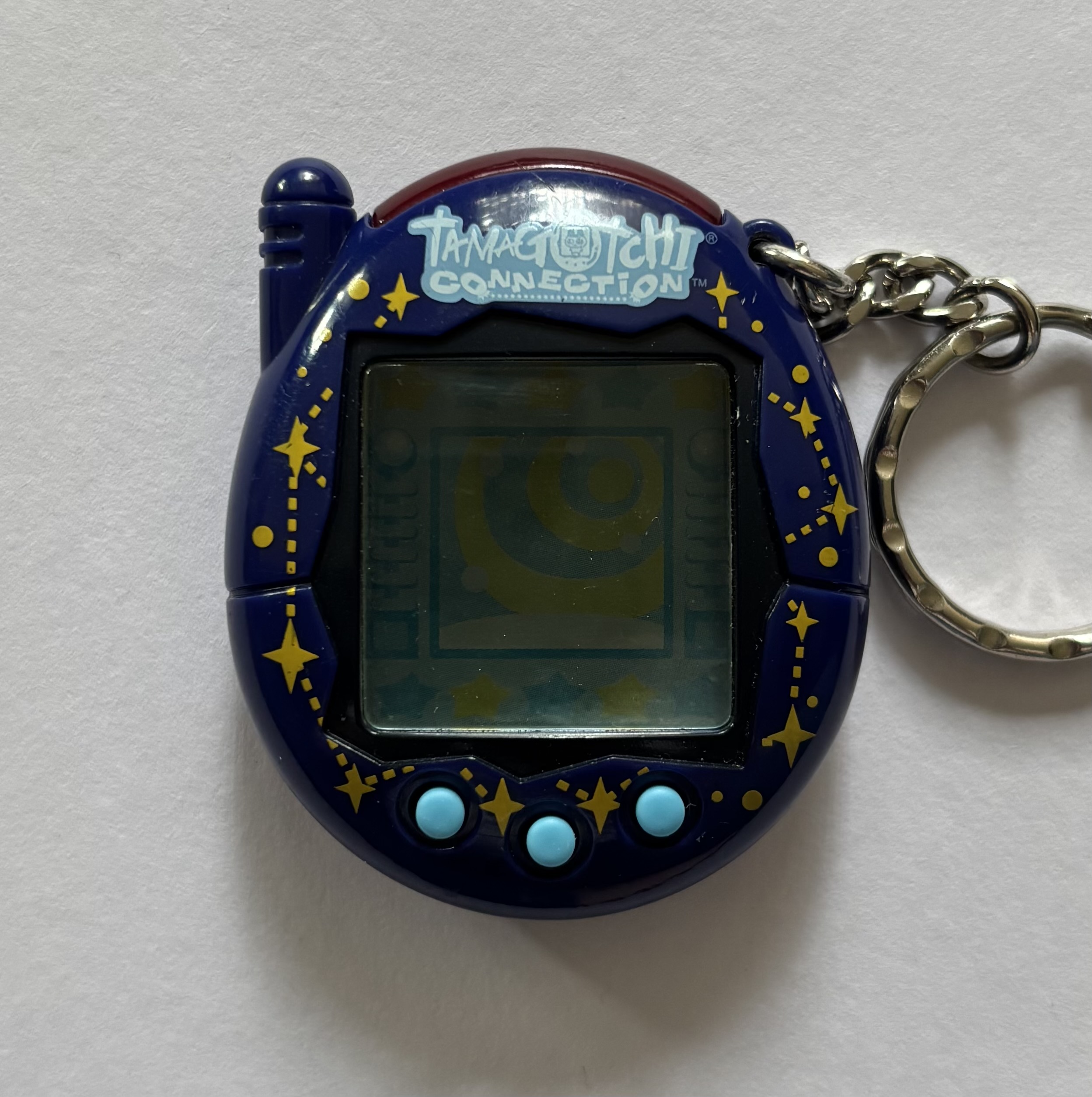 Dark blue shell. Black screen border. Light blue buttons. Yellow stars connected by dashed lines surrounding screen. 'Tamagotchi Connection' label on light blue background.